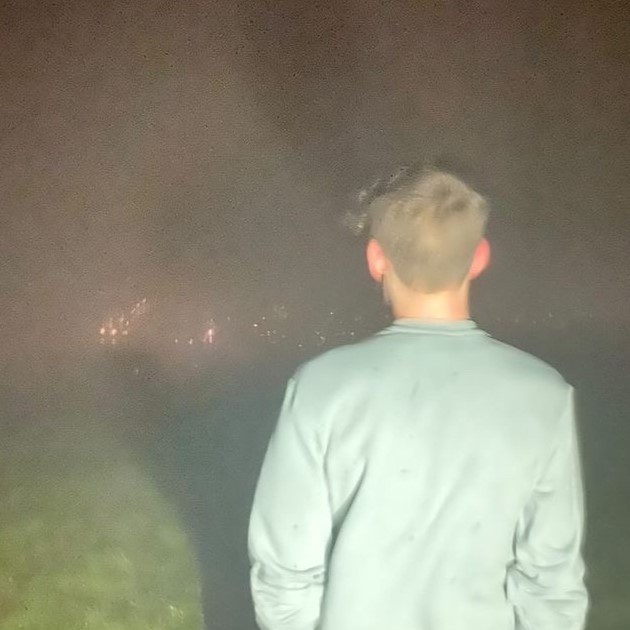 A dimly lit image of the back of Linus Sewell, wearing a turquoise sweater and jeans.